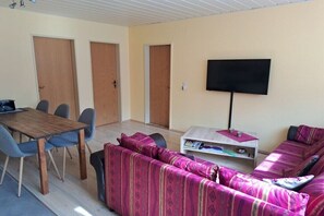 Ein Zimmer mit Esstisch aus Holz, blauen Stühlen, einem pinkfarbenen Sofà mit Muster sowie einem Fernseher an der Wand und einem kleinen weißen Beistelltisch.