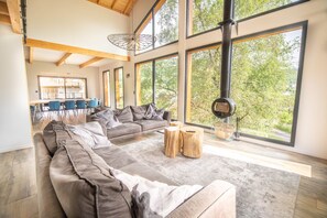 Un salon moderne doté d’un grand canapé d’angle, d’un coin repas avec des chaises bleues et de grandes fenêtres offrant une vue sur les arbres.