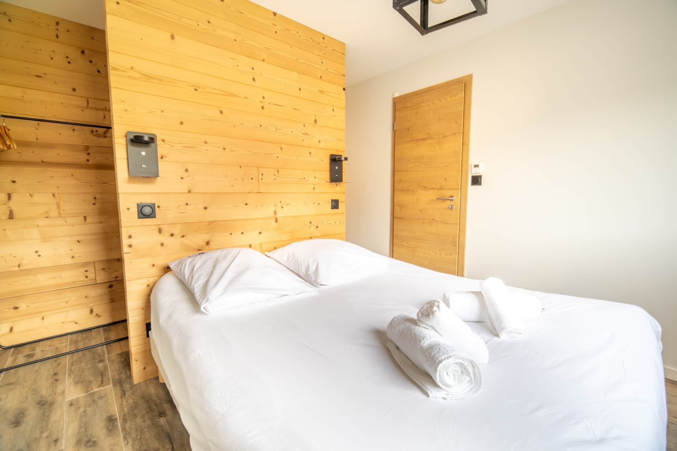 A hotel room with a wooden wall, a bed with white linens, a wooden door, and a wall-mounted light fixture.