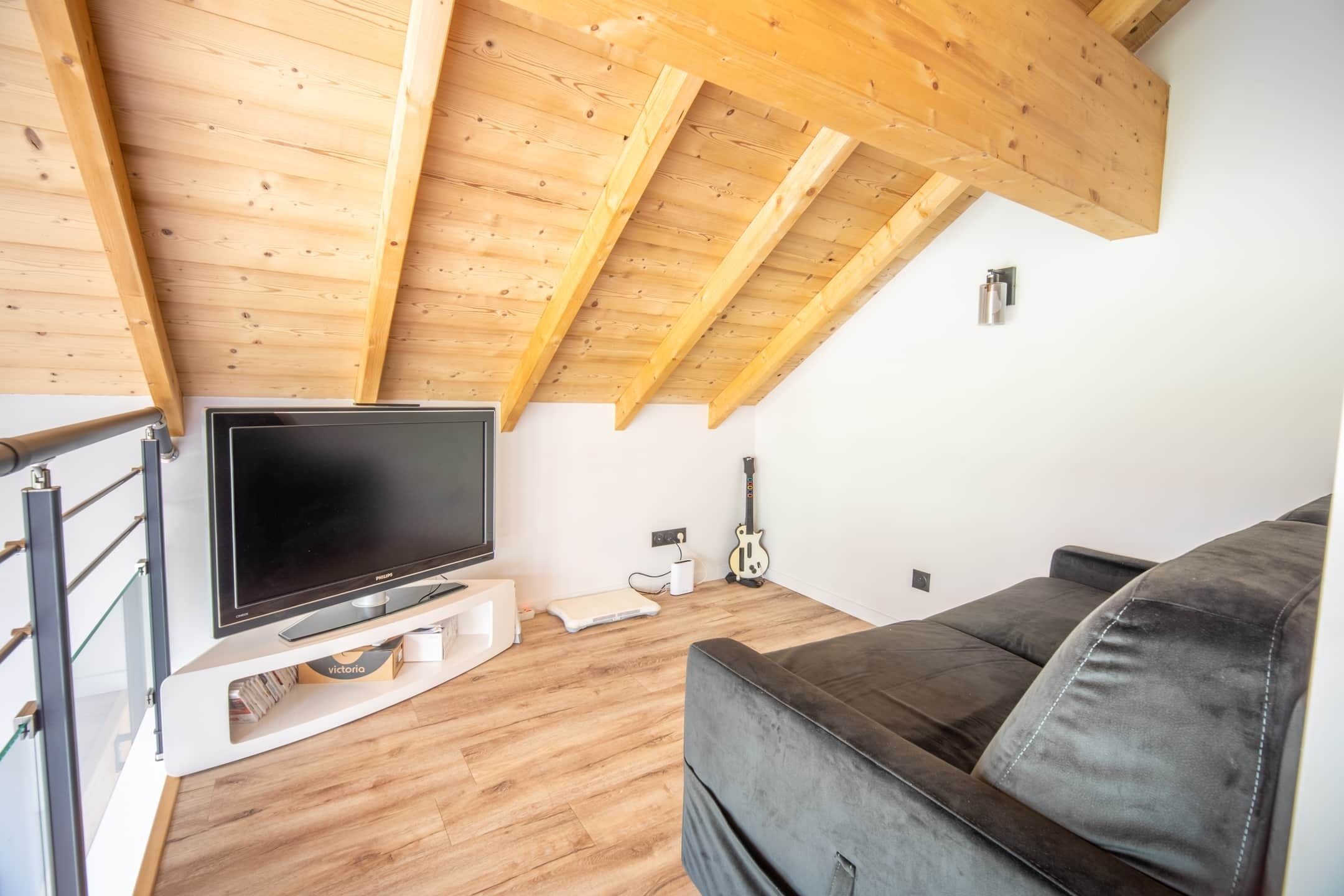 Una sala de estar moderna con techo de madera, un televisor de pantalla plana sobre un mueble blanco, un sofá de cuero negro y una guitarra apoyada en la pared.