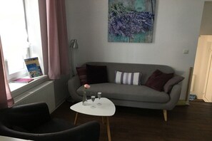 Ein Wohnzimmer mit grauem Sofa, einem kleinen weißen Tisch mit Vase und Gläsern, einem Fenster mit rosa Vorhängen und einem Bild an der Wand.