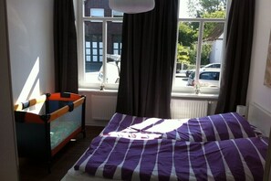 Ein Zimmer mit einem Bett, einem kleinen Tisch, einem Fenster mit Vorhängen und Blick auf geparkte Autos.