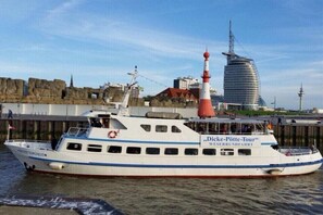 Ein Boot namens 'Dicke-Potte-Tour' mit weißem Rumpf und rotem Schornstein liegt an einem Steg. Im Hintergrund ist eine felsige Küstenlinie und ein modernes Gebäude zu sehen.