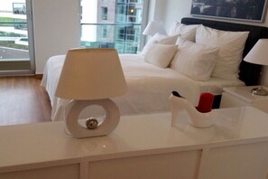 Quarto de hotel com cama, mesa de cabeceira com abajur, porta-copos e vista para edifícios.