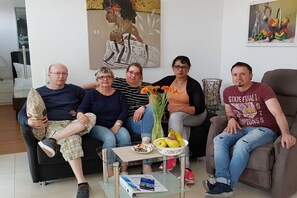 Eine Gruppe von Menschen sitzt auf einer Couch in einem Wohnzimmer, inklusive einem Glastisch, einer Vase mit Blumen und einem Gemälde an der Wand.