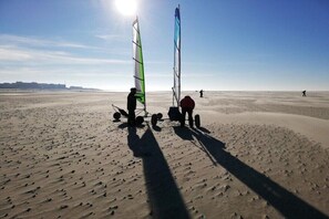 Ein sandiger Strand mit zwei Windsurfbrettern und Segeln, einem klaren Himmel und Gebäuden in der Ferne.
