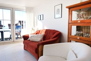 Ein Wohnzimmer mit einem roten Sofa, einem weißen Sessel, einer Hollape mit Gläsern und Blick durch eine Glastür.