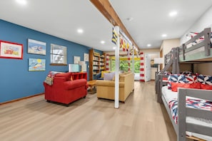 A cozy living room with a red sofa, a beige sofa, and a bunk bed with colorful bedding. There are bookshelves, a wooden coffee table, and a window with curtains. - As You Like It | Steps to the Beach | Peek-a-boo Views (Santa Rosa Beach)