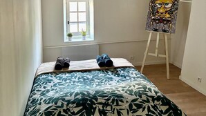 Ein Schlafzimmer mit Bett, Fenster, einem Gemälde auf einem Staffelei und einer kleinen Pflanze auf dem Fensterbrett.