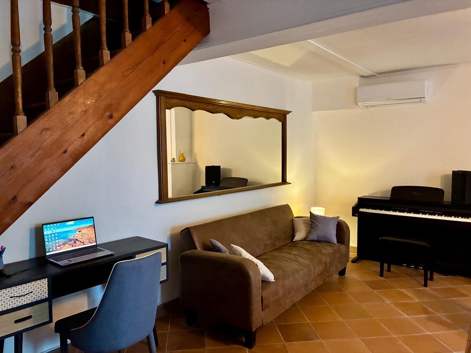 Un soggiorno con una scala, un divano, una scrivania con un portatile, un pianoforte e un grande specchio.