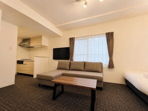 Ein Hotelzimmer mit einer Couch, einem kleinen Tisch, einem Fernseher, einem Bett, einer Küchenzeile und einem Fenster mit Vorhängen.