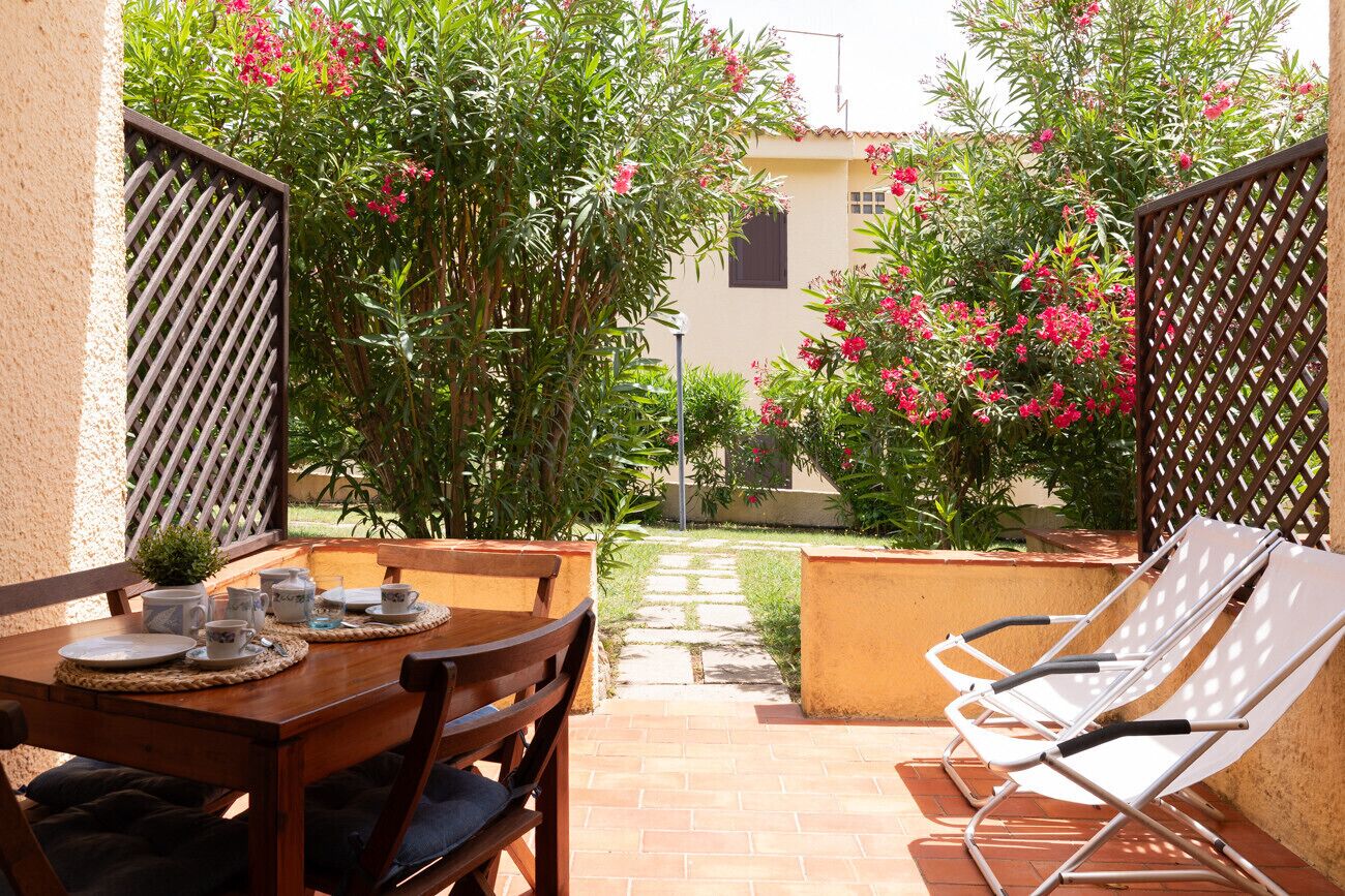 Un patio con un tavolo apparecchiato per due persone, due poltrone relax e un giardino con piante fiorite.