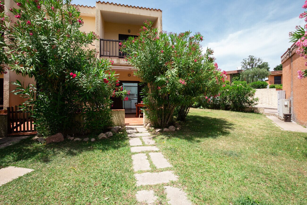 A residential building with a stone pathway leading to the entrance, surrounded by lush greenery and flowering plants.