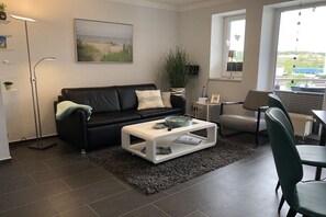 A modern living room with a black leather sofa, a white coffee table, a green armchair, and a view of the outdoors through a large window. - More about the Wadden Sea Doris Frenzel (Cuxhaven)