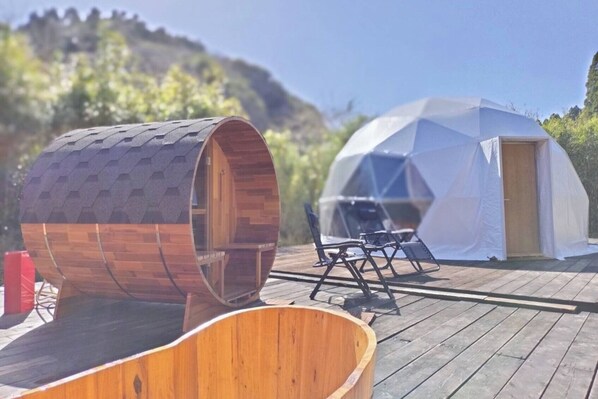 A wooden barrel-shaped structure and a geodesic dome tent on a wooden deck with outdoor furniture.