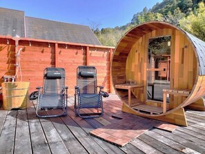 Una estructura tipo pod de madera con un banco en su interior, dos sillones reclinables en el exterior y una terraza de madera.
