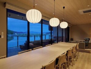 Un espace de salle à manger moderne avec une longue table et des chaises, de grandes fenêtres offrant une vue sur un plan d’eau et des montagnes, et des luminaires suspendus.