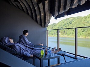 Eine Person entspannt sich in einer Liege am Fenster mit Blick auf einen See, ein Tisch steht vor ihr.