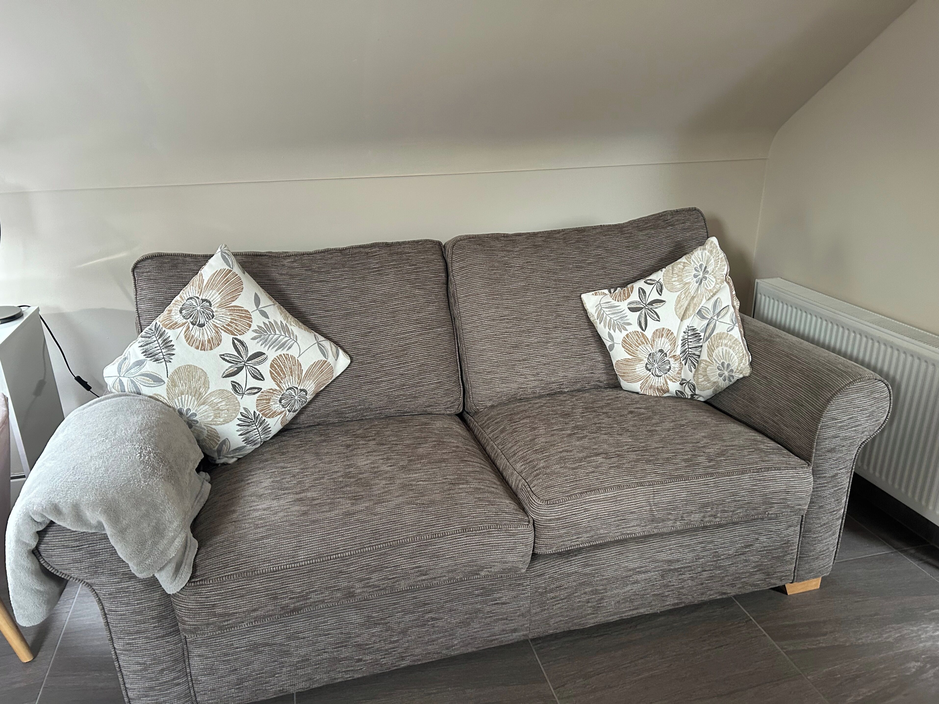 A grey sofa with floral-patterned cushions in a room with a radiator and a tiled floor.