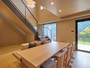 Un intérieur moderne avec une table à manger en bois, des bancs, un escalier et une grande fenêtre donnant sur un paysage verdoyant.