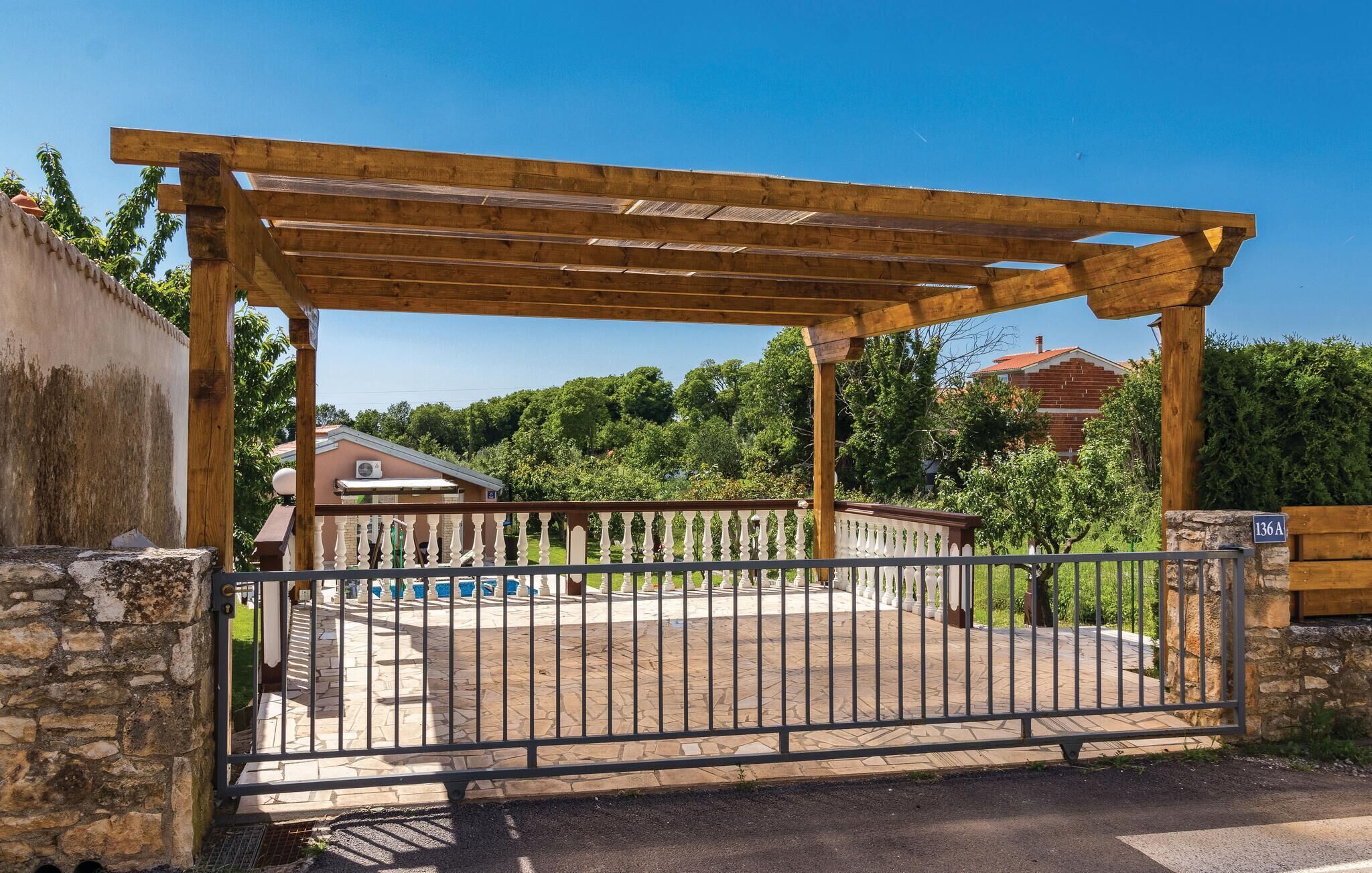 A gated entrance with a wooden pergola, stone walls, and a clear view of a residential area with trees and a house in the background.