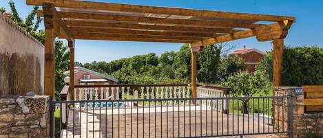 A gated entrance with a wooden pergola, stone walls, and a clear view of a residential area with trees and a house in the background.