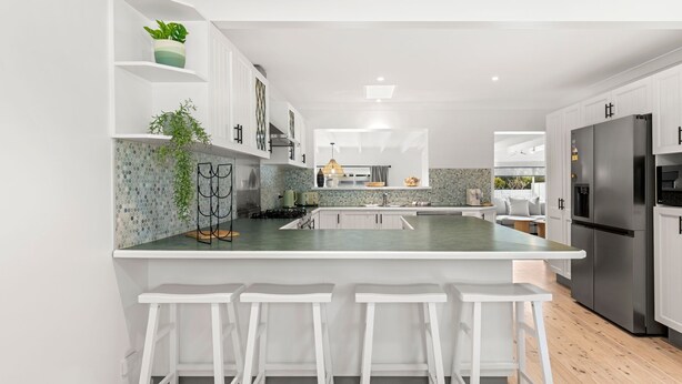 A modern kitchen with white cabinetry, a central island with bar stools, a stainless steel refrigerator, and a tiled backsplash. - Samphire House - Fireplace with Heated Swimming Pool (Callala Bay)