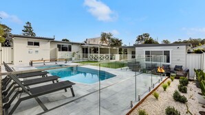 A modern pool area with a glass-enclosed deck, lounge chairs, and a fire feature. - Samphire House - Fireplace with Heated Swimming Pool (Callala Bay)