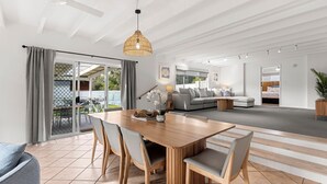 A spacious living area with a dining table, chairs, a sofa, and a large window. - Samphire House - Fireplace with Heated Swimming Pool (Callala Bay)
