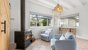 A modern living room with a wood-burning stove, two blue armchairs, a wooden cabinet, and large windows. - Samphire House - Fireplace with Heated Swimming Pool (Callala Bay)