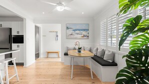 A modern kitchen with white cabinetry, a dining area with a bench and table, and a large window with tropical plants. - Samphire House - Fireplace with Heated Swimming Pool (Callala Bay)