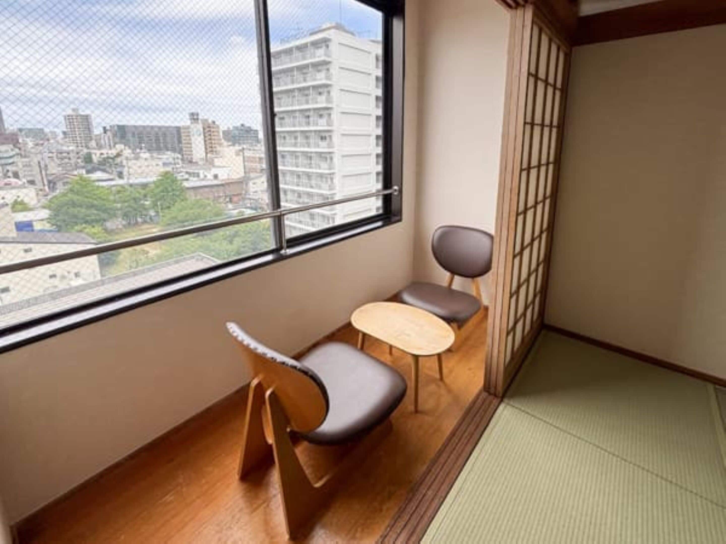 Una habitación con vista a un paisaje urbano, suelo de tatami, una mesa de madera baja y dos sillas.