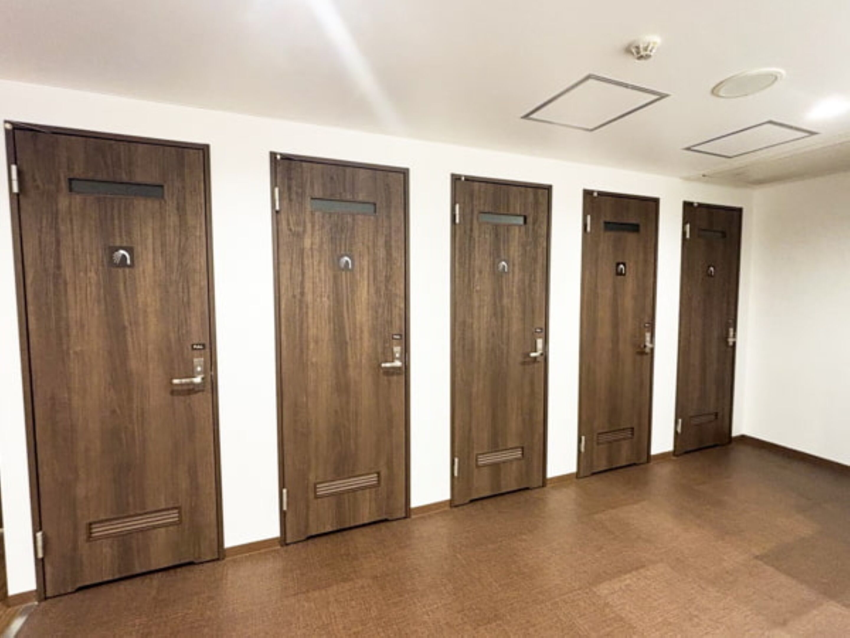 A row of wooden doors with a handle and a vent, white walls, and a tiled floor.