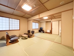 A tatami mat room with tatami mats, a low table, two chairs, a television, and a sliding door.