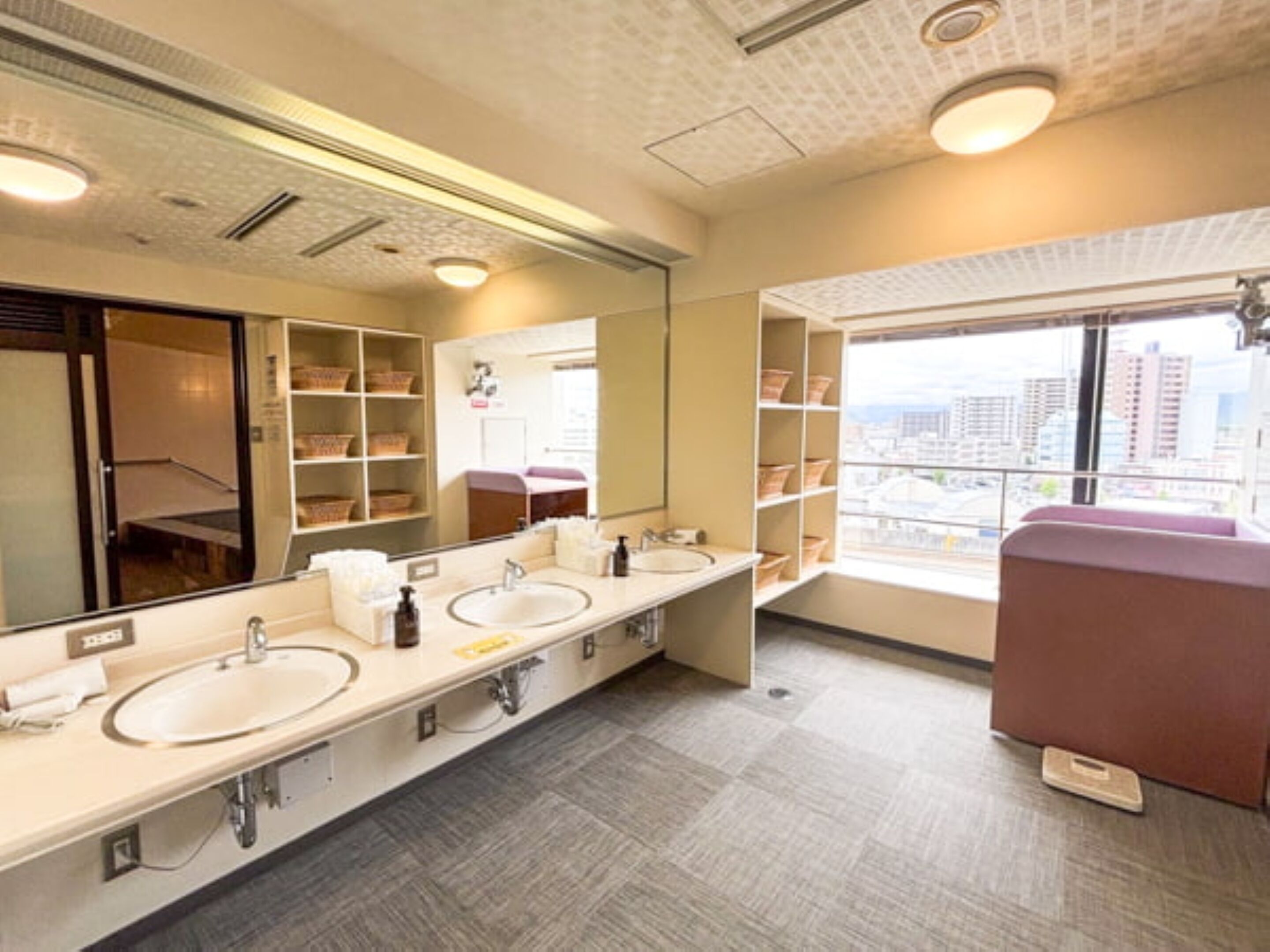 Un baño moderno con un espejo grande, dos lavamanos, repisas con artículos de aseo y una ventana con vista a la ciudad.