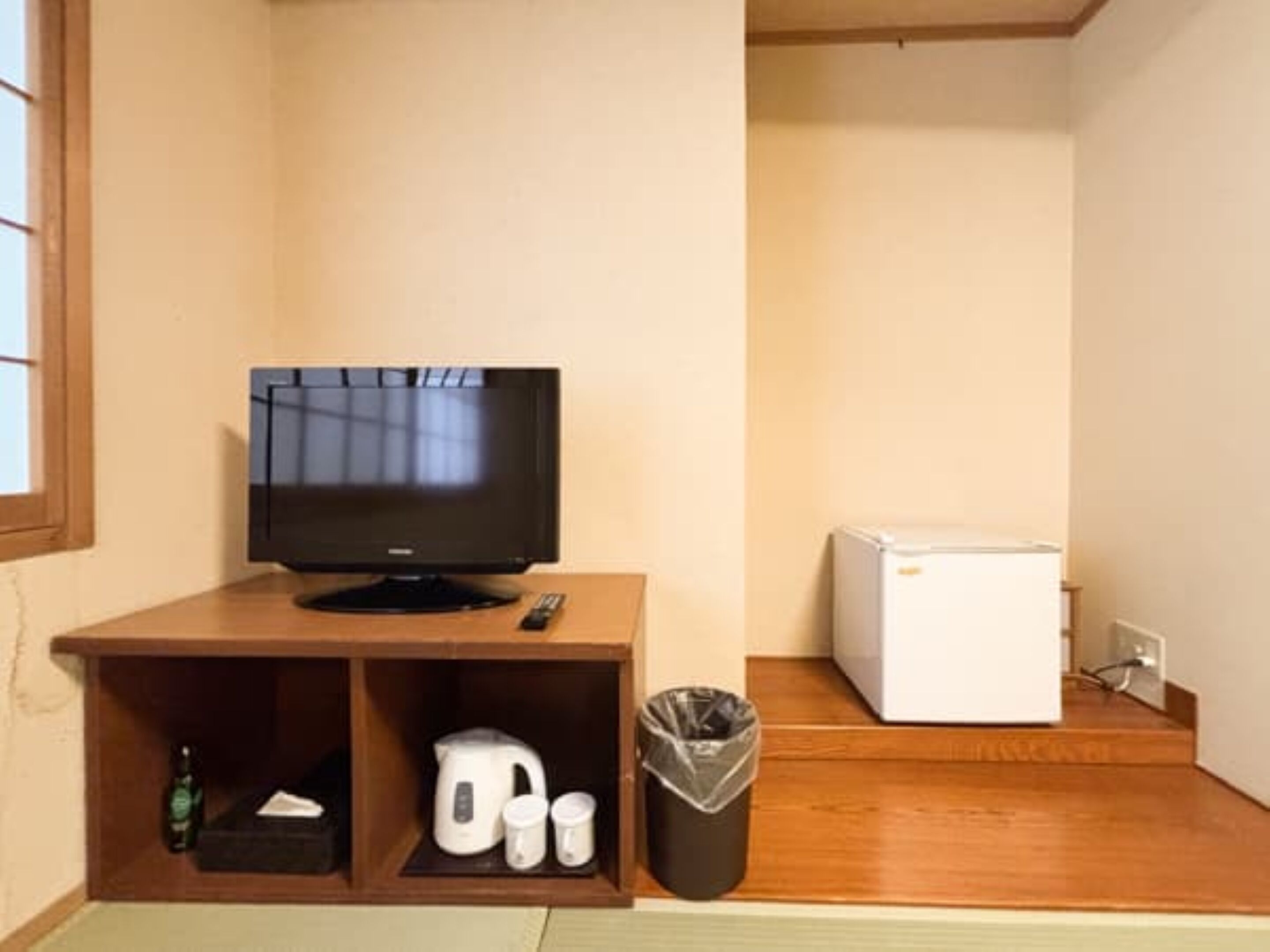 A small room with a television on a wooden stand, a white refrigerator, a trash bin, and a shelf with a kettle and cups.