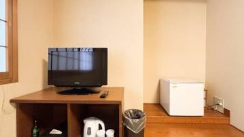 A small room with a television on a wooden stand, a white refrigerator, a trash bin, and a shelf with a kettle and cups.
