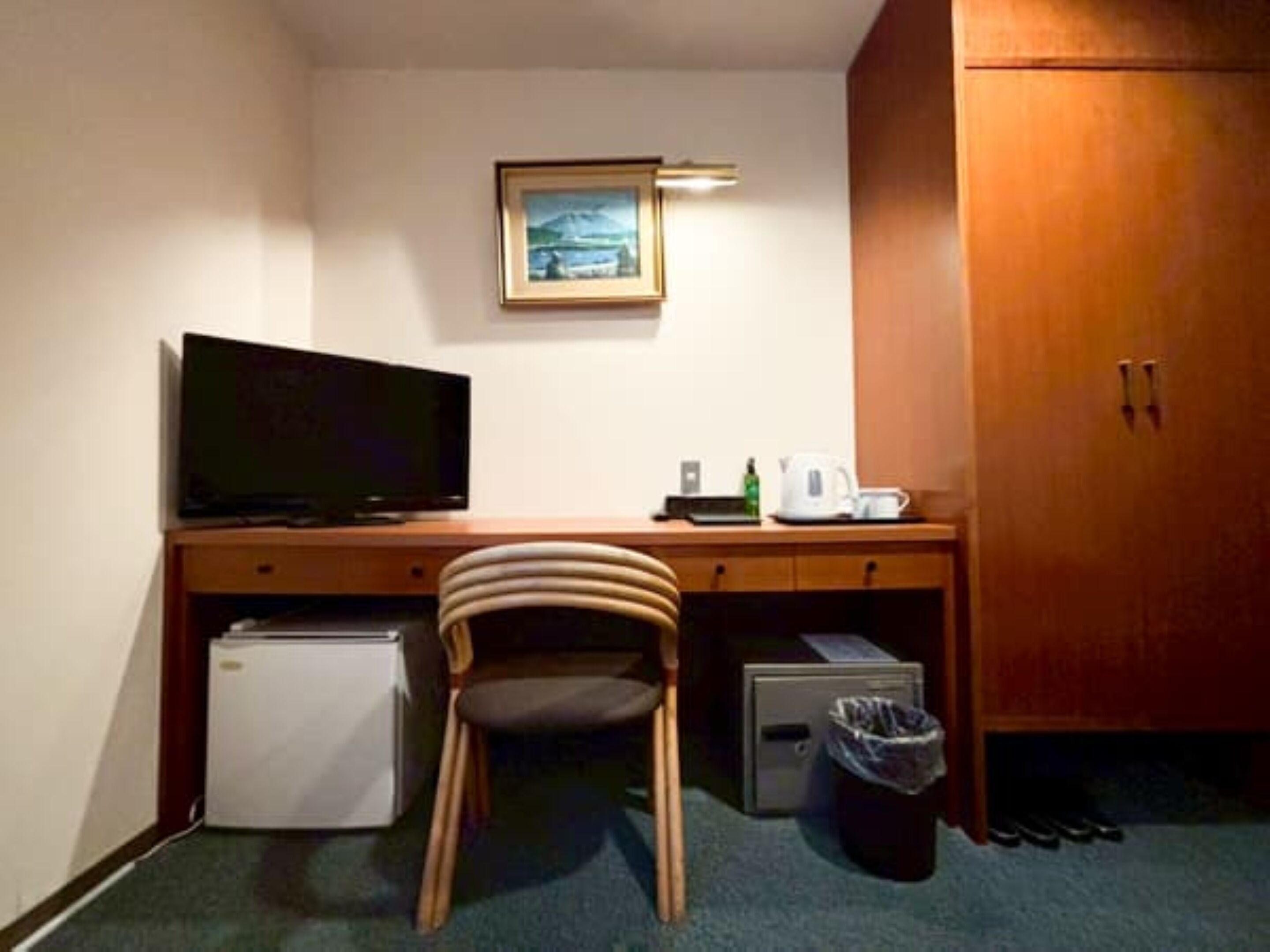 Ein kleines Zimmer mit Schreibtisch, Fernseher, Kühlschrank, Stuhl, Mülleimer und einem Holzschrank.