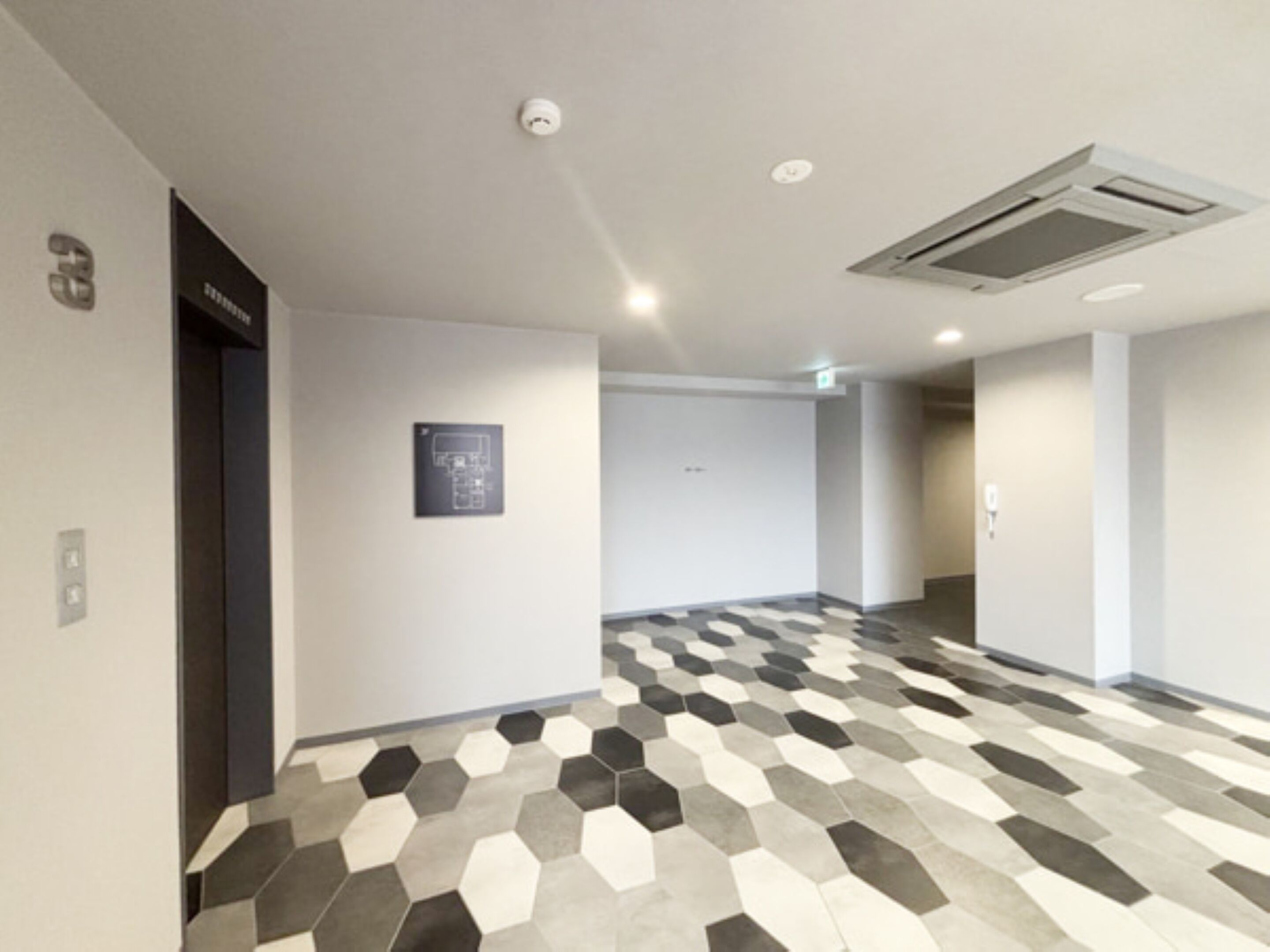 A hallway with a geometric patterned floor, a door with a sign, and a wall-mounted light fixture.