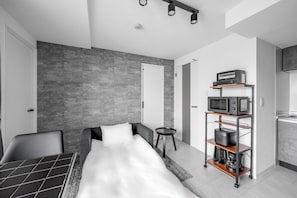 Ein modernes Hotelzimmer mit einem Bett, einem kleinen Tisch, einer Mikrowelle, einer Kaffeemaschine und einem Regal mit Küchengeräten.
