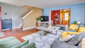 A living room with a sofa, coffee table, and TV. There is a staircase and a kitchen area visible.