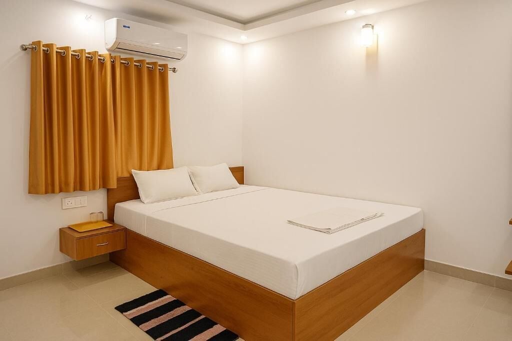 A modern bedroom with a wooden bed frame, white bedding, a nightstand, a lamp, and an air conditioning unit.