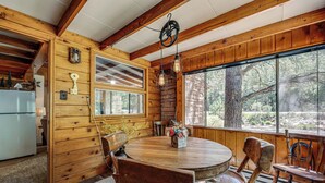 Interior de una cabaña de madera con una mesa redonda de comedor, sillas de madera, un refrigerador y ventanas grandes que dan a un bosque.