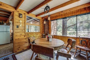 Ein Holzblockhaus-Interieur mit einem runden Esstisch, Holzstühlen, einem Kühlschrank und großen Fenstern mit Blick auf den Wald.
