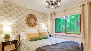 Een slaapkamer met een bed, nachtkastje, lamp, spiegel en een raam met uitzicht op bomen.