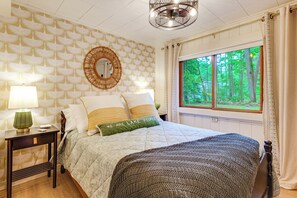 A bedroom with a bed, a nightstand, a lamp, a mirror, and a window with a view of trees. - Waterfront Tee Lake Cabin: Dock, Fire Pit + More! (Lewiston)