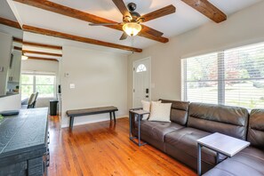 Ein Wohnzimmer mit braunem Ledersofa, einem kleinen Tisch, einem Deckenventilator, Holzbalken und einem Fenster mit Jalousien.