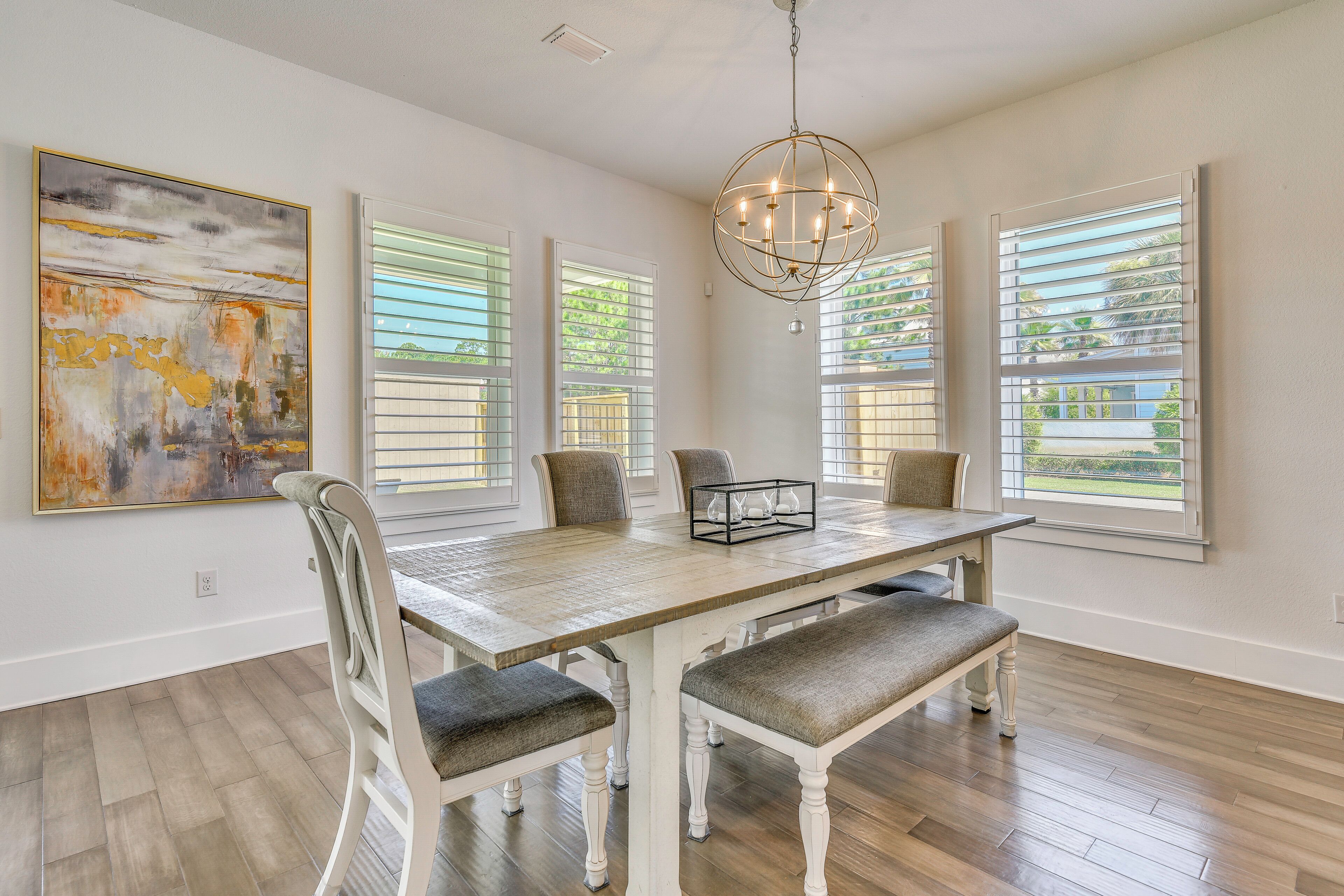 Une salle à manger avec une table en bois, des chaises blanches, un lustre et de grandes fenêtres équipées de stores.