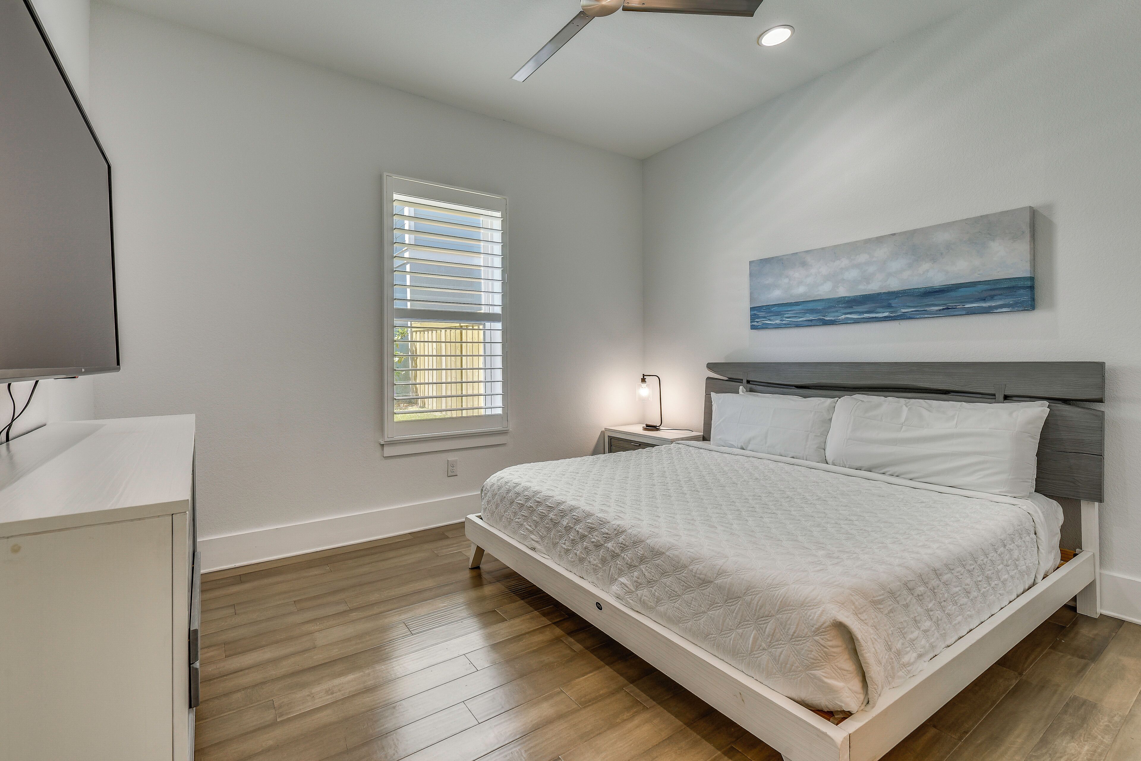 A bedroom with a bed, bedside table, lamp, artwork, and a window.