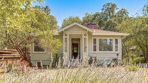 Une maison de plain-pied avec un porche à l’avant, entourée d’un jardin où poussent de hautes herbes et des arbres.